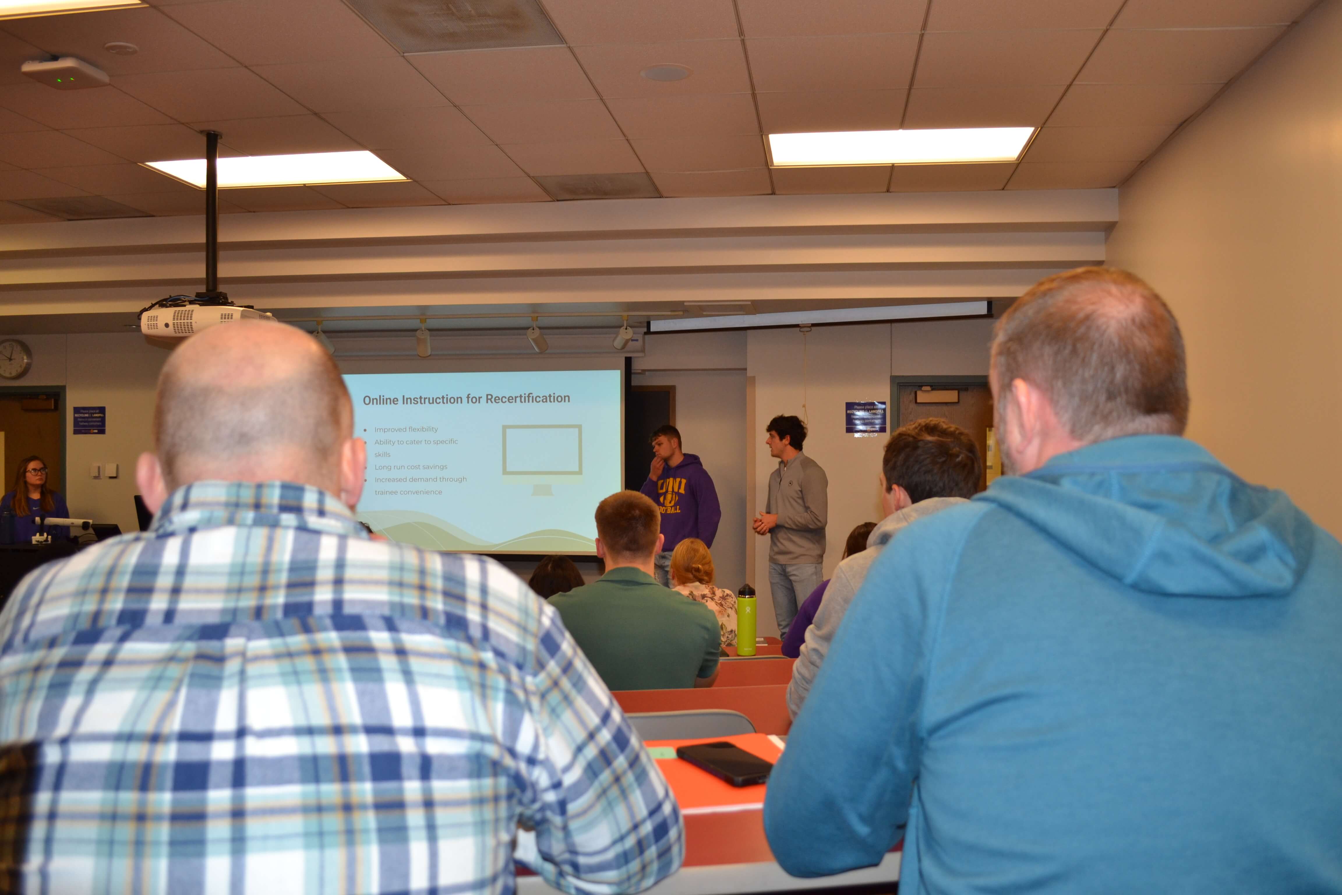 Backs of two men sitting in the back of a classroom watching student presentations