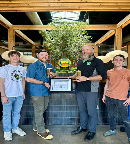 four men in front of large tree holding a plaque and glass of beer