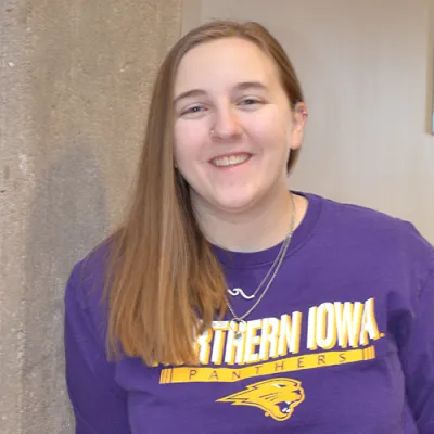 Link, Young woman wearing a University of Northern Iowa purple shirt leaning against a cement pillar, profile page